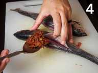 A hand is holding the slit open on a plastic chopping board and stuffing it with sambal using a spoon. A hand is holding the slit open on a plastic chopping board and stuffing it with sambal using a spoon.