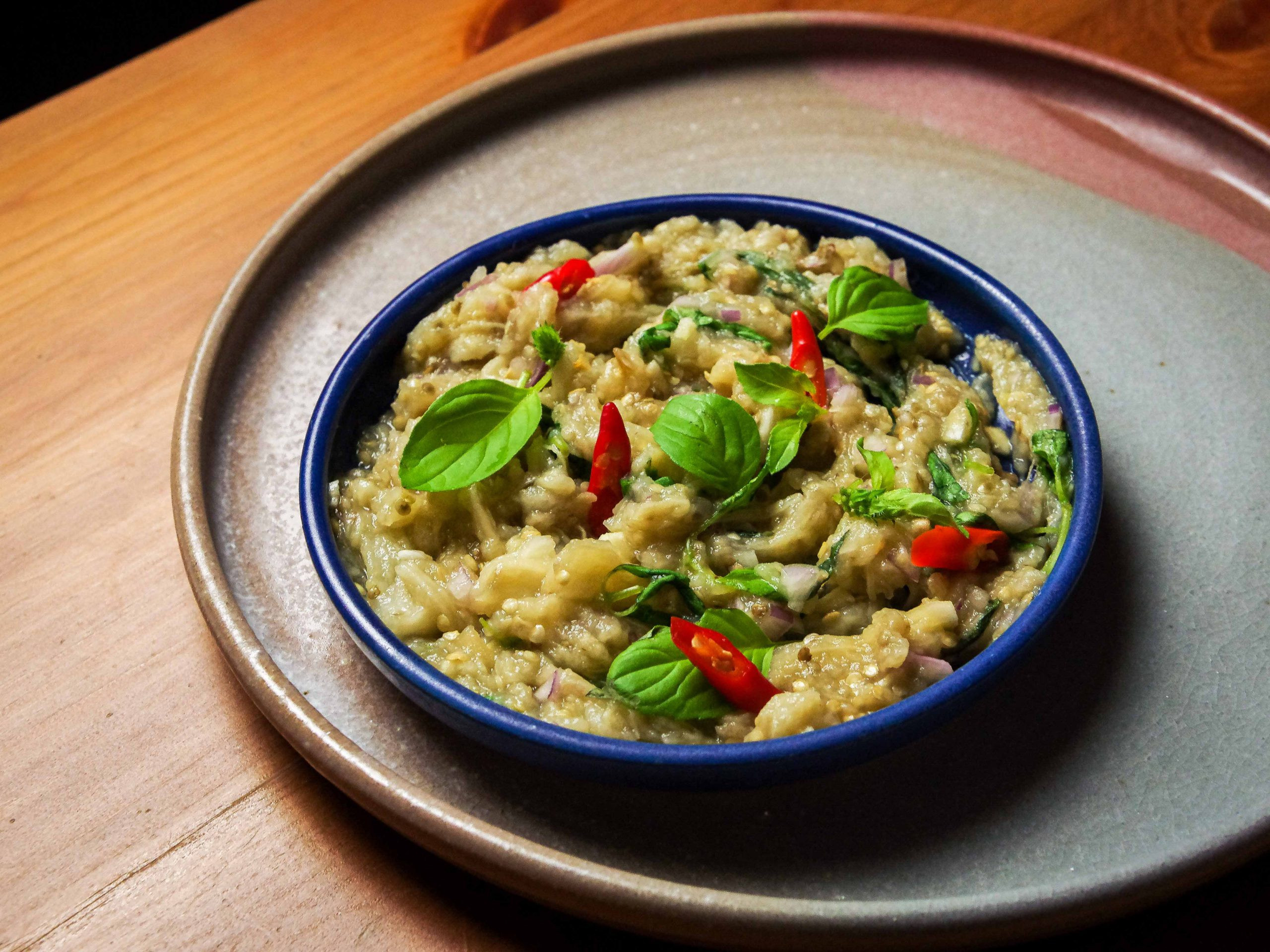 Mashed roasted eggplant sits in a small blue plate, decorated with lemon basil leaves. The small plate is sitting on a grey-pink plate, on a wooden table.