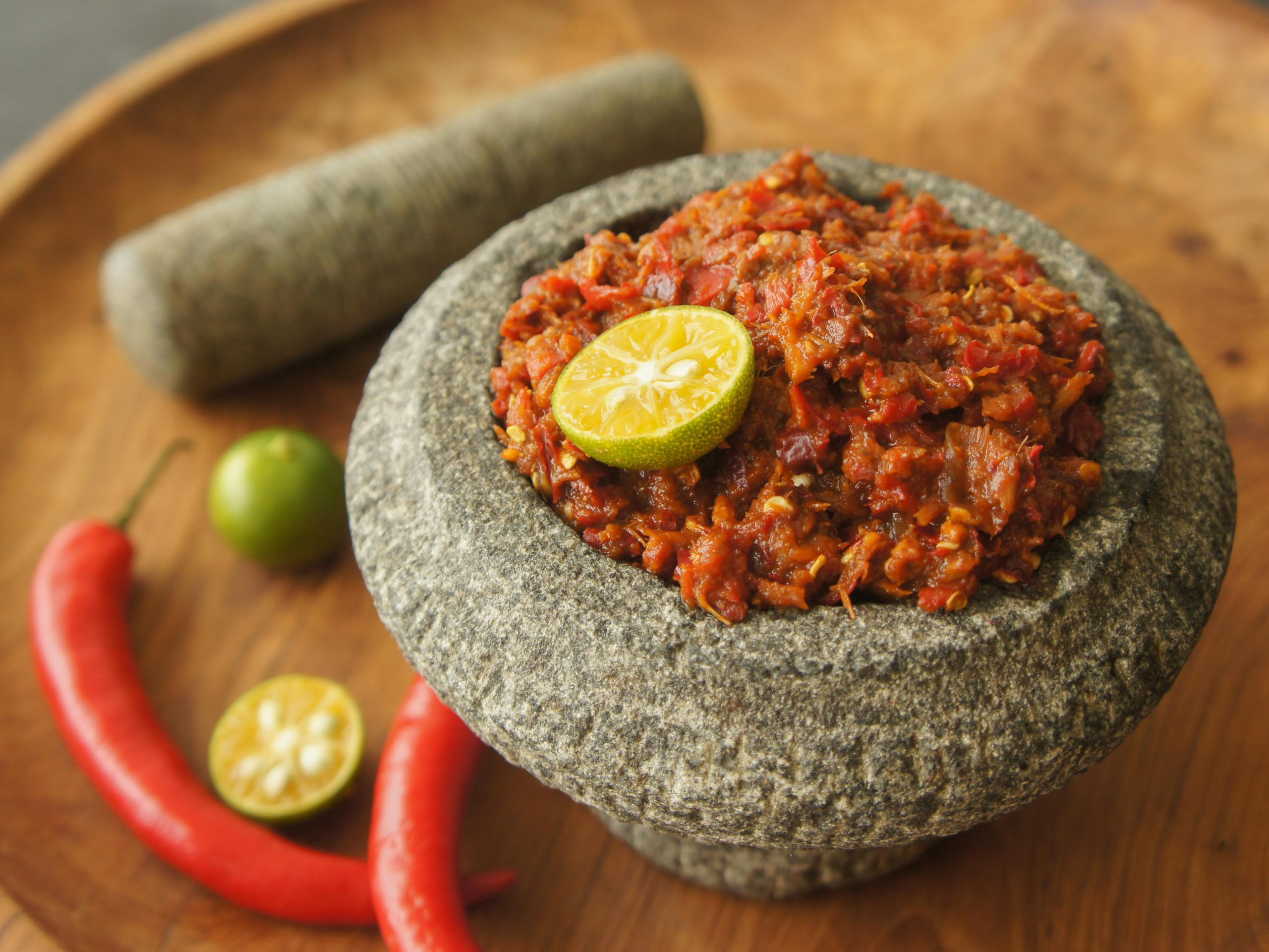 Sambal belacan vegan di dalam lesung batu, bersama anak lesung, beberapa cili dan potongan limau di atas latar belakang kayu.