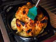 A green pastry brush is basting a whole roast chicken, that is in a cast iron pan with cabbage slices. A green pastry brush is basting a whole roast chicken, that is in a cast iron pan with cabbage slices.