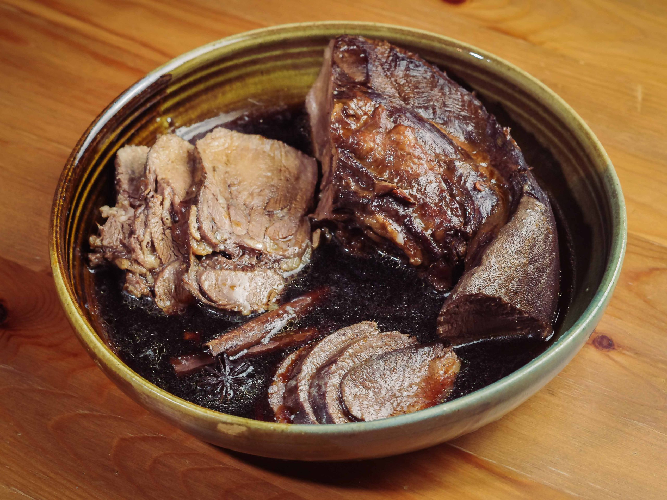 Slices of braised beef tongue are nestled in a green-brown bowl, on a wooden table.