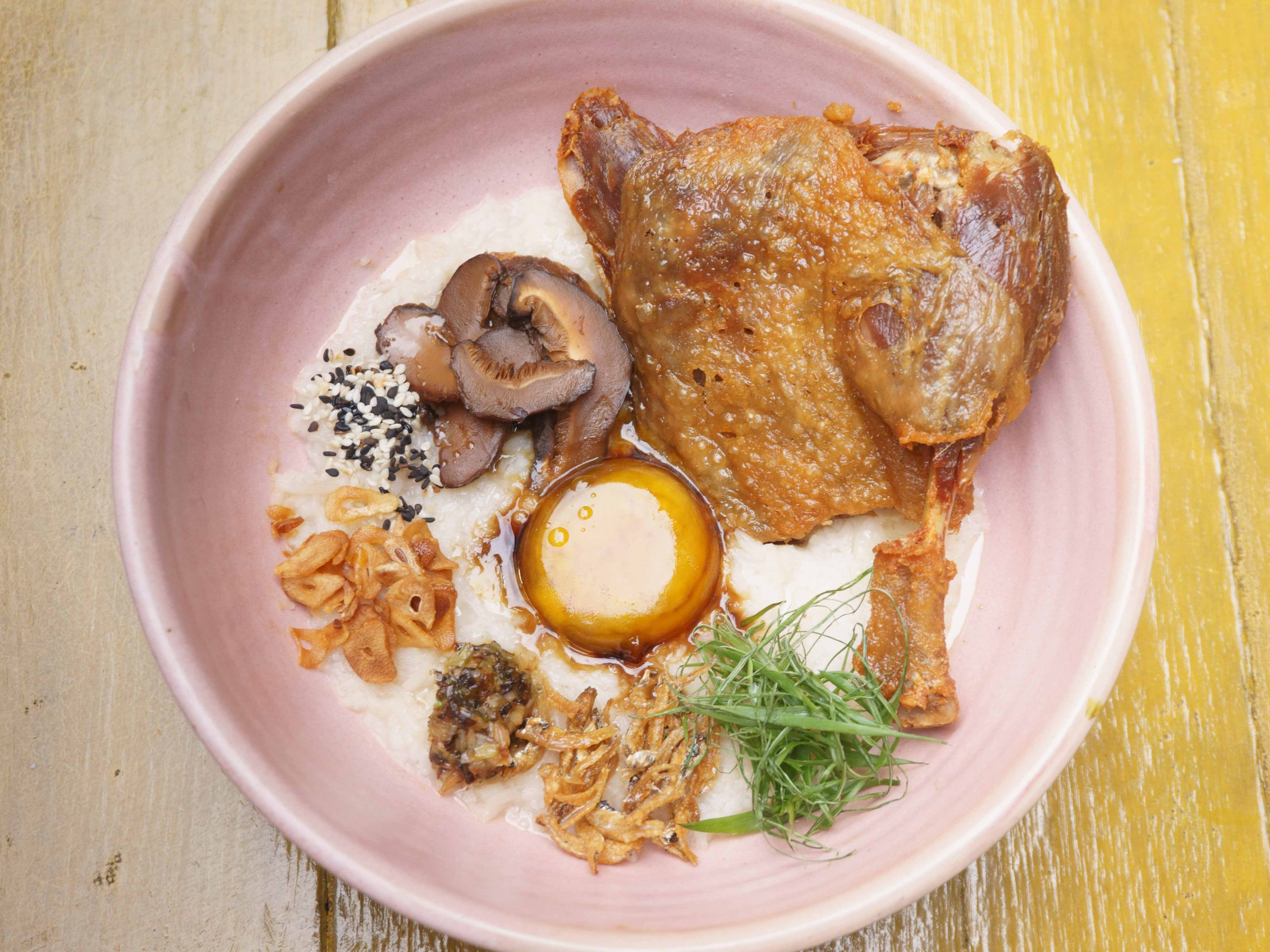 Bubur nasi dengan pelbagai lauk termasuk confit itik, di dalam mangkuk merah jambu di atas meja kayu.
