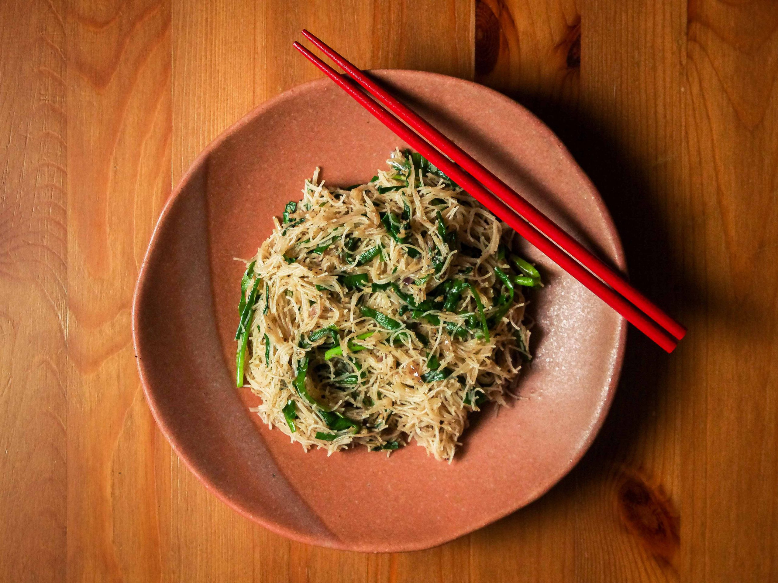 Fried meehoon and kucai sit on a terracotta coloured plate, on a wooden table. A pair of red chopsticks lay diagonally on the rim of the plate.