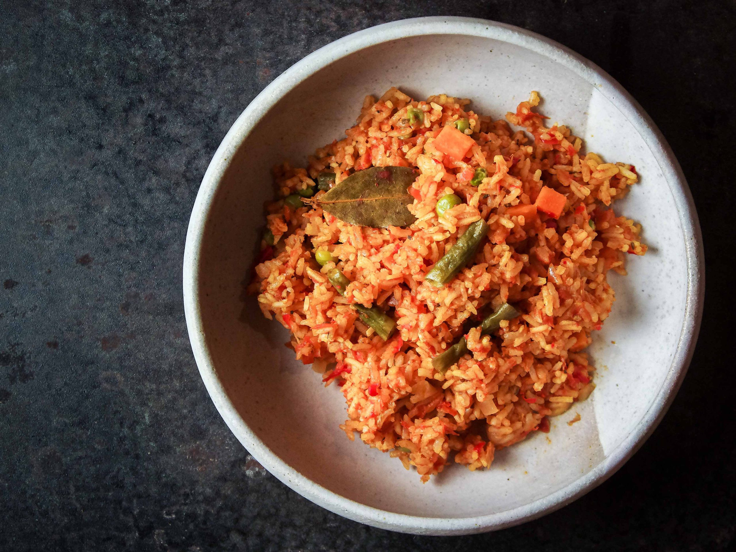 Tomato-coloured rice is served in a grey plate, studded with vegetables. The background is dark grey steel.